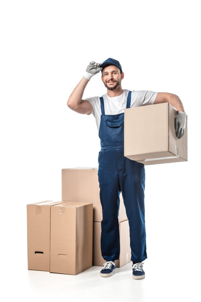 handsome mover in uniform adjusting hat and looking at camera while carrying cardboard box isolated